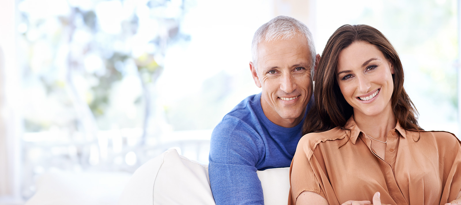 An elderly couple sitting on a couch, smiling and looking at the camera.