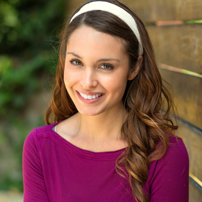 The image shows a smiling woman with long hair, wearing a purple top and a headband, standing against a wooden fence in a sunny outdoor setting.