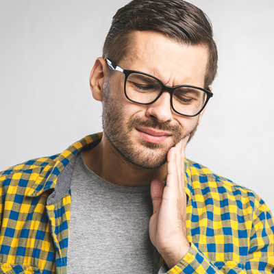 Man with beard and glasses, wearing a yellow plaid shirt, holding his hand to his mouth in a thoughtful or pensive pose.