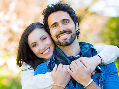 Two adults, one male and one female, are embracing in a warm hug against an outdoor backdrop.