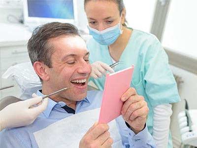 The image shows a man sitting in a dental chair, holding up a pink card with a smile, while a dental professional is attending to him.