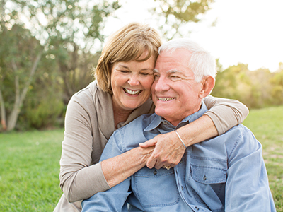 An elderly couple embracing, smiling and looking at the camera outdoors during sunset.