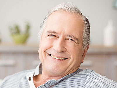 The image shows an older man with gray hair, wearing a blue shirt and smiling at the camera. He appears to be seated in a home environment.