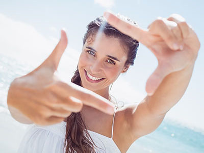 This image shows a smiling woman with long hair, wearing a white top, taking a selfie by holding up her hand with fingers extended to frame the shot. She is outdoors with a clear sky and ocean in the background.